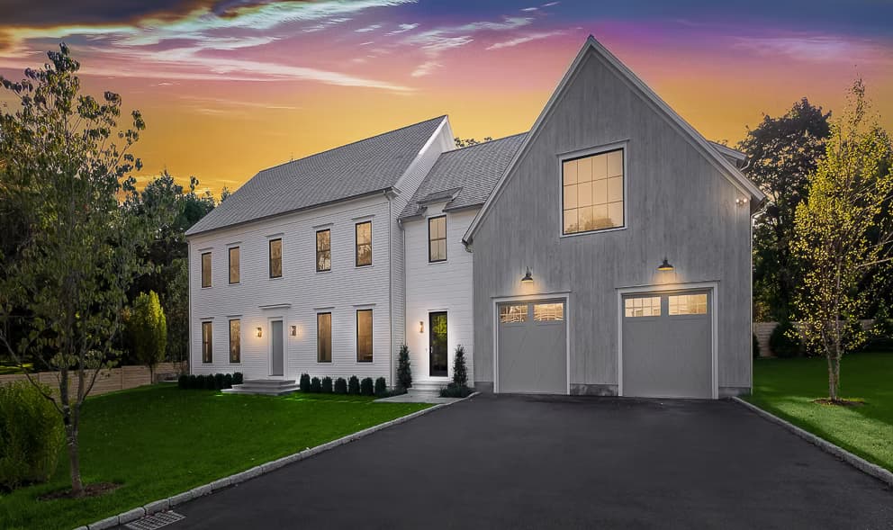 A New England colonial home at dusk with lit windows, glowing wall sconces, and carriage lighting
